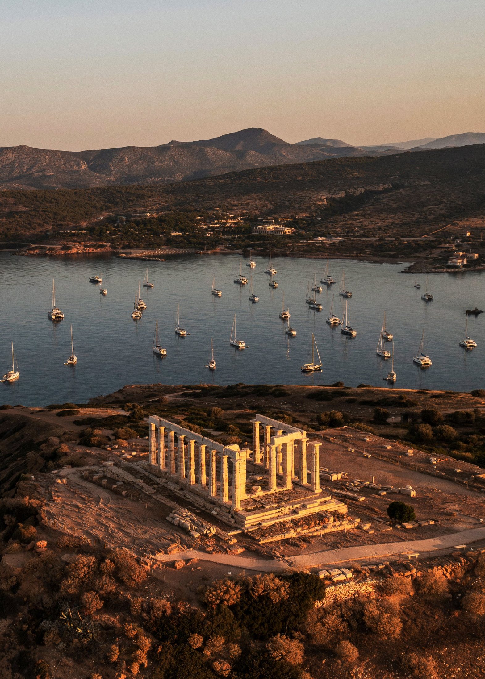 A temple on a headland in low evening sun, with boats at anchor in the bay behind.