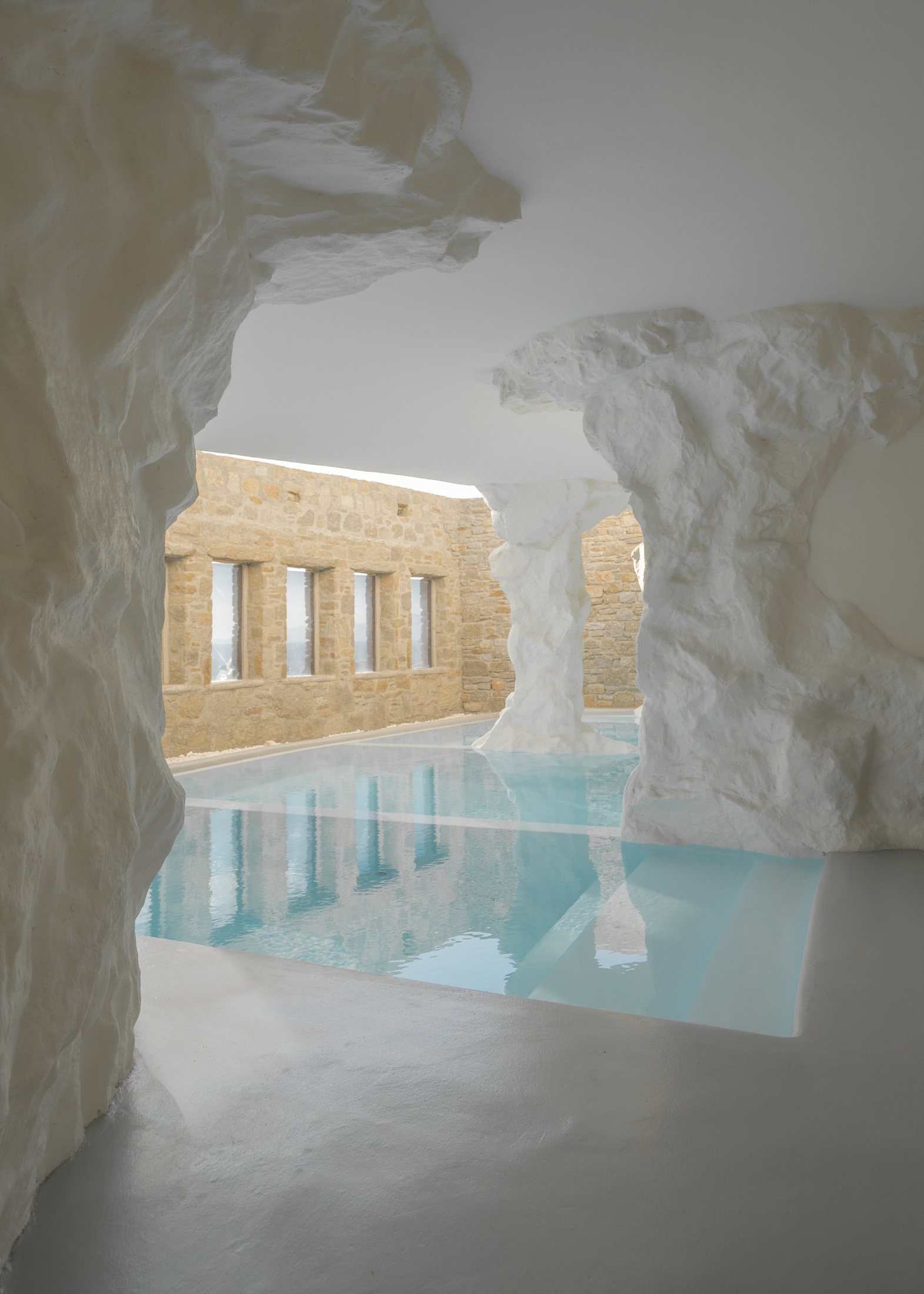 An indoor pool between white plastered rock forms, with a stone wall and windows reflected in the water.